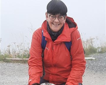 Eine Person in einer roten Jacke sitzt auf einem Stein. Hinter ihr ist ein nebliger Hintergrund mit Pflanzen.