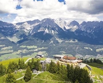 Eine beeindruckende Berglandschaft mit hohen Gipfeln und grünen Wiesen. Im Vordergrund sind Bäume und Gebäude zu sehen, die die natürliche Schönheit der Umgebung ergänzen.