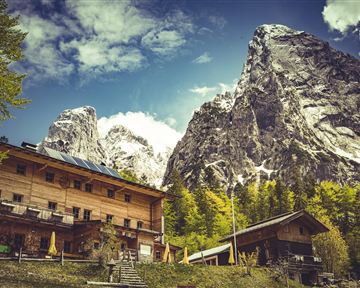 A picturesque mountain landscape with tall peaks and a charming wooden lodge in the foreground. The sky is partly cloudy and the nature is lush green.