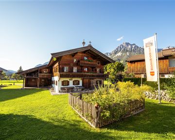 Ein traditionelles alpenländisches Holzhaus mit einem liebevoll gestalteten Garten. Im Hintergrund sind majestätische Berge und ein klarer blauer Himmel zu sehen.