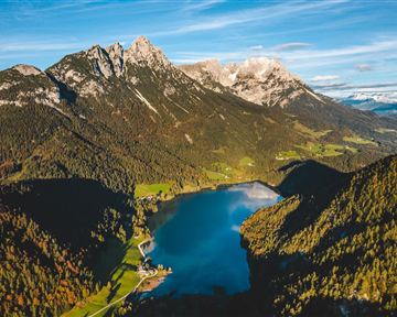 Eine atemberaubende Berglandschaft mit schneebedeckten Gipfeln und einem klaren See im Vordergrund. Die Natur ist umgeben von grünen Wäldern und Feldern.