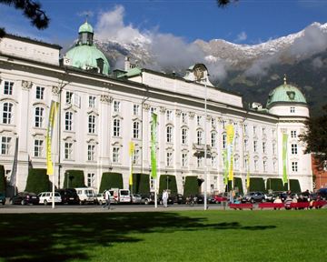 Ein imposantes Gebäude mit grünen Kuppeln und einer schönen Fassade. Im Vordergrund gibt es eine grüne Wiese und die umliegenden Berge sind teilweise schneebedeckt.