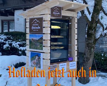A small outdoor sales booth for local products stands in a snowy environment. The booth displays signs with the name "Käserei Ackerhube" and information about the offerings.