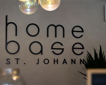 A modern sign with the inscription "home base St. Johann" on a wall. In the background, a plant and a warm light source can be seen.