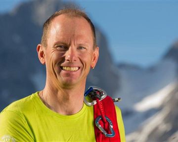 Ein lächelnder Mann in einem leuchtend gelben Shirt steht vor einer bergigen Landschaft. Er trägt Kletterausrüstung und strahlt Abenteuerlust aus.