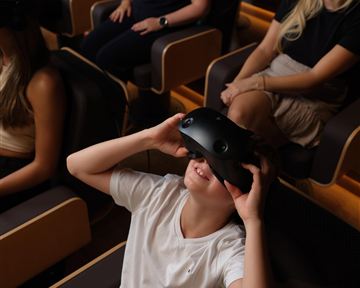 Eine Gruppe von Personen sitzt in bequemen Sesseln und trägt Virtual-Reality-Headsets. Ein Kind lächelt und genießt die Erfahrung.