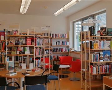 A bright library with shelves full of books. In the middle are tables and chairs for readers.