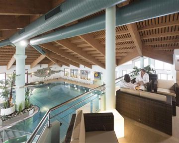 Ein eleganter Innenpool mit Holzdecke und Pflanzen. Im Vordergrund sitzen zwei Personen auf einer gemütlichen Sitzgruppe.