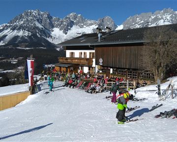 An alpine ski landscape with snow-covered mountains and a wooden house in the background. Several skiers and snowboarders are enjoying the slope.