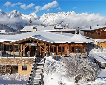 A rustic mountain cabin in the snow with a stunning view of the surrounding mountains. The sky is clear and the landscape is characterized by a beautiful winter atmosphere.