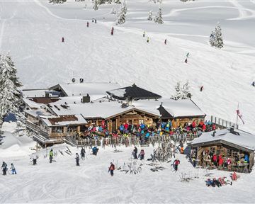 Eine gemütliche Skihütte umgeben von Schnee und Bergen. Viele Menschen genießen die Winterlandschaft und das Skifahren.