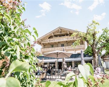 A picturesque building with a balcony, surrounded by green plants and a beautiful sky. In the foreground, tables and chairs are visible outdoors.