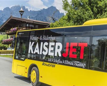 A yellow bus with the inscription "KaiserJET" drives through a picturesque landscape. In the background, mountains and a cozy alpine hut can be seen.