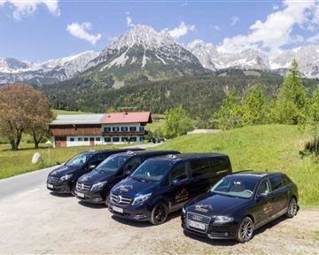 Eine Gruppe von Fahrzeugen steht vor einem malerischen Bergpanorama. Im Hintergrund sind grüne Wiesen und schneebedeckte Gipfel zu sehen.