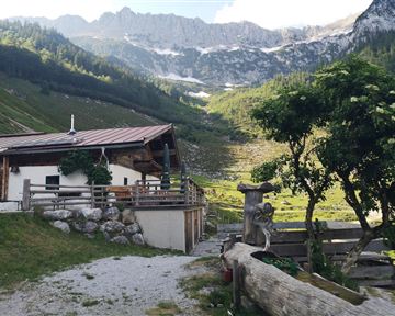 A cozy cabin in the middle of a picturesque mountain landscape. In the background, green meadows and high mountains can be seen.