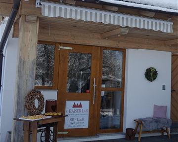 Ein gemütlicher Eingang zu einem Holzhaus mit einem Schild von "Kaiser Lidl". Vor der Tür steht ein Tisch mit Gebäck und eine Sitzgelegenheit.