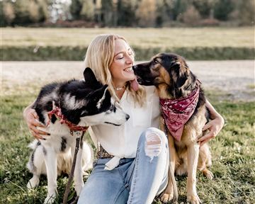 A woman is sitting on the grass and embracing two dogs. She smiles happily while the dogs are lovingly by her side.