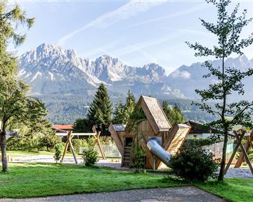 Eine Spielplatzlandschaft mit einem kreativen Holzspielgerät und Schaukeln, umgeben von grünen Wiesen. Im Hintergrund erheben sich majestätische Berge unter einem klaren Himmel.