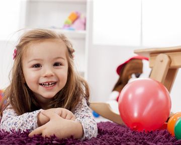 A cheerful girl is lying on a purple carpet and smiling. Colorful toys and a red ball are around her.