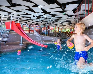 Ein Kinderbecken mit einer roten Rutsche. Zwei Kinder spielen im Wasser und spritzen fröhlich umher.