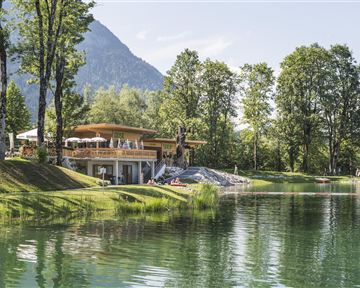 A picturesque building on the shore of a clear lake, surrounded by lush trees. In the background, gentle mountains can be seen, creating an idyllic atmosphere.