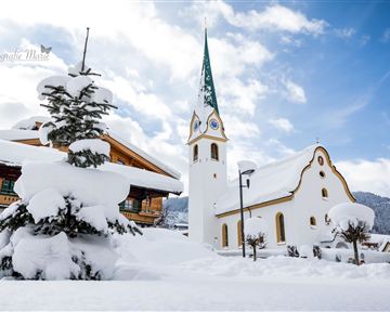 Ein verschneites Dorf mit einer schönen Kirche und einem hohen, grünen Kirchturm. Die Umgebung ist ruhig und winterlich, bedeckt mit frischem Schnee.