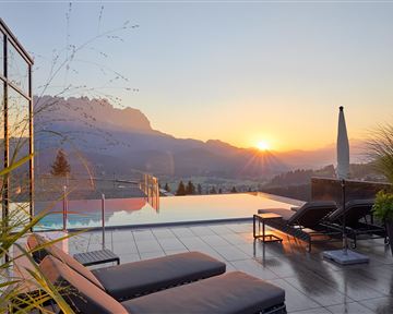 Ein wunderschöner Poolbereich mit Liegen und Blick auf die Berge. Die Sonne geht hinter den Bergen unter und taucht die Szene in warmes Licht.