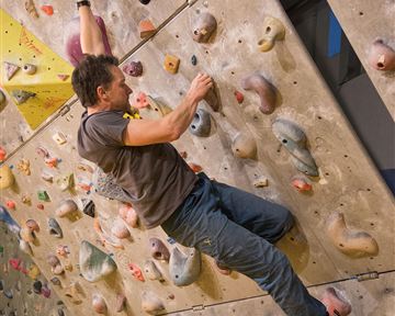 Ein Kletterer, der an einer Kletterwand mit verschiedenen Griffen unterwegs ist. Die Wand ist bunt und hat zahlreiche kleine Haltegriffe.