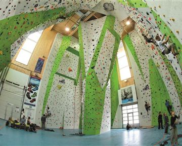 Eine Kletterhalle mit hohen Wänden und bunten Klettergriffen. Menschen klettern an den Wänden, während andere zuschauen und sich vorbereiten.