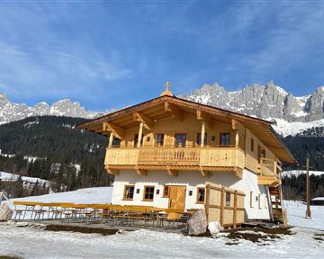 A beautiful wooden house in the mountains, surrounded by snow. The clear sky and the majestic mountains in the background give the landscape a picturesque atmosphere.