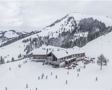 A picturesque ski resort with a wooden house and snow-covered mountains. Many people are on the slopes.