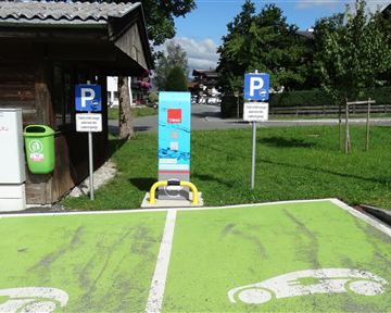 A parking lot for electric vehicles with a charging station and visible markings. In the background, there are additional parking spaces and a green area.