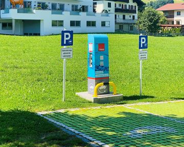 A parking lot with a ticket machine is located on a green meadow. In the background, residential buildings can be seen.