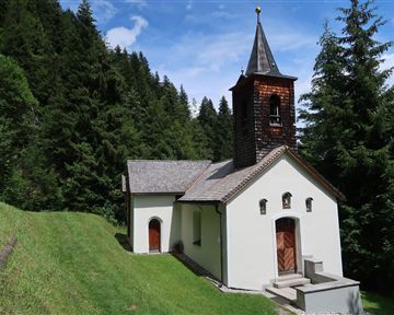 Eine kleine Kirche umgeben von dichten Bäumen. Der Himmel ist blau mit einigen Wolken.
