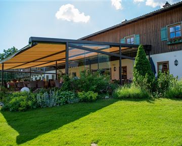 Ein gemütliches Haus mit einer großen Terrasse und einem hellen Sonnensegel. Der Garten ist grün und üppig, mit vielen Pflanzen und Sitzgelegenheiten.