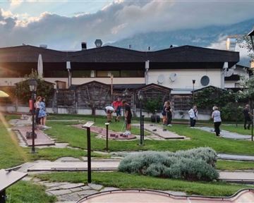 A mini-golf facility with multiple players outdoors. Trees and a building are visible in the background.