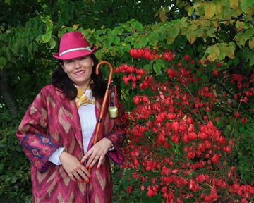 Eine lächelnde Frau in einem bunten Outfit steht neben einem blühenden Strauch mit roten Früchten. Sie trägt einen Hut und hält einen Gehstock in der Hand.