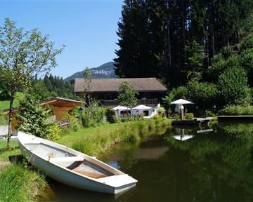 A calm pond with a boat and surrounding green meadows. In the background, a wooden house and some sun umbrellas can be seen.