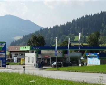 A OMV gas station surrounded by mountains and trees. The sky is clear and the landscape is green.