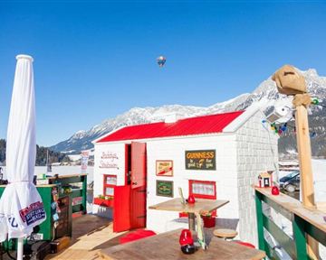 A cozy wooden house with a red roof in a snow-covered mountain landscape. The sky is clear blue and various lights adorn the area.
