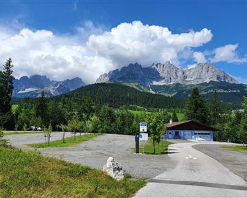 A picturesque parking lot with a view of majestic mountains and a green landscape. The sky is partly cloudy and blue.