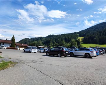 A parking lot with several cars and a beautiful mountain landscape in the background. The sky is clear with some clouds.