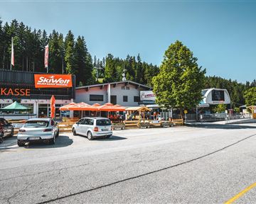 Ein Parkplatz vor einem modernen Ski Resort mit mehreren Geschäften und Restaurants. Die Umgebung ist von Bäumen umgeben und es herrscht schönes Wetter.