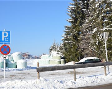 Eine verschneite Landschaft mit einem Parkplatzschild und einem Verbotsschild. Im Hintergrund sind große grüne Ballen und schneebedeckte Bäume zu sehen.