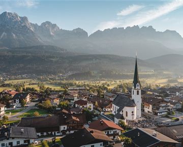 A picturesque Alpine landscape with a charming village view. In the foreground stands a church with a pointed tower, surrounded by mountains and meadows.