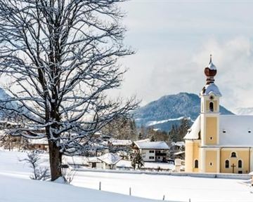 Eine malerische Schneelandschaft mit einer gelben Kirche und einem großen Baum. Die Berge im Hintergrund runden das winterliche Bild ab.