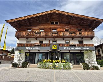 Eine moderne Bankfiliale im alpinen Stil mit Holzverkleidung. Vor der Bank stehen gelbe Fahnen und es gibt eine grüne Bepflanzung.