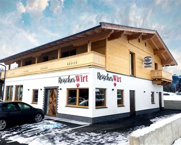 A modern building with wooden cladding and a bright outdoor area. The inscription "Reischer Wirt" is clearly visible on the facade.