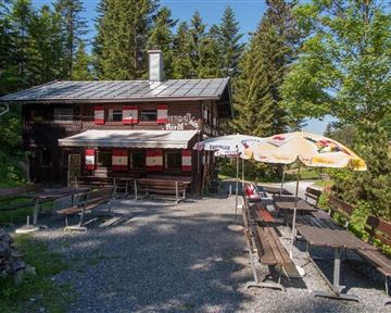 Eine gemütliche Berghütte umgeben von Bäumen. Vor der Hütte stehen Bänke und Tische, ideal für eine Pause in der Natur.