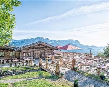 A cozy mountain cabin with traditional wooden design and a beautiful terrace. In the background, majestic mountains and a blue sky are visible.